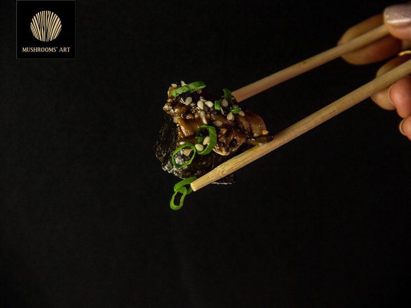 Chopsticks που κρατούν ένα ρολό από φύλλα nori και από πάνω έχουν King Oyster μανιτάρια σε μαύρο φόντο